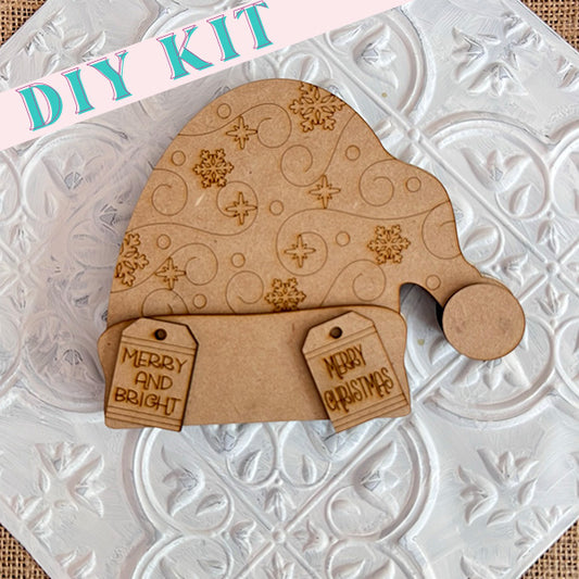 This DIY kit from Janet's Craft Corner features a chunky Santa Hat wooden cutout embellished with intricate snowflake engravings. It comes with two delightful tags reading "Merry and Bright" and "Merry Christmas," making it an ideal addition to your holiday home decor against the textured white background.