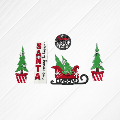 Janet's Craft Corner Santa Interchangeable Porch Set is a DIY seasonal home decor kit with 5 pieces, including potted Christmas trees, festive signs like "SANTA is coming to town," a red sleigh, and an unfinished "Santa STOP here" sign.