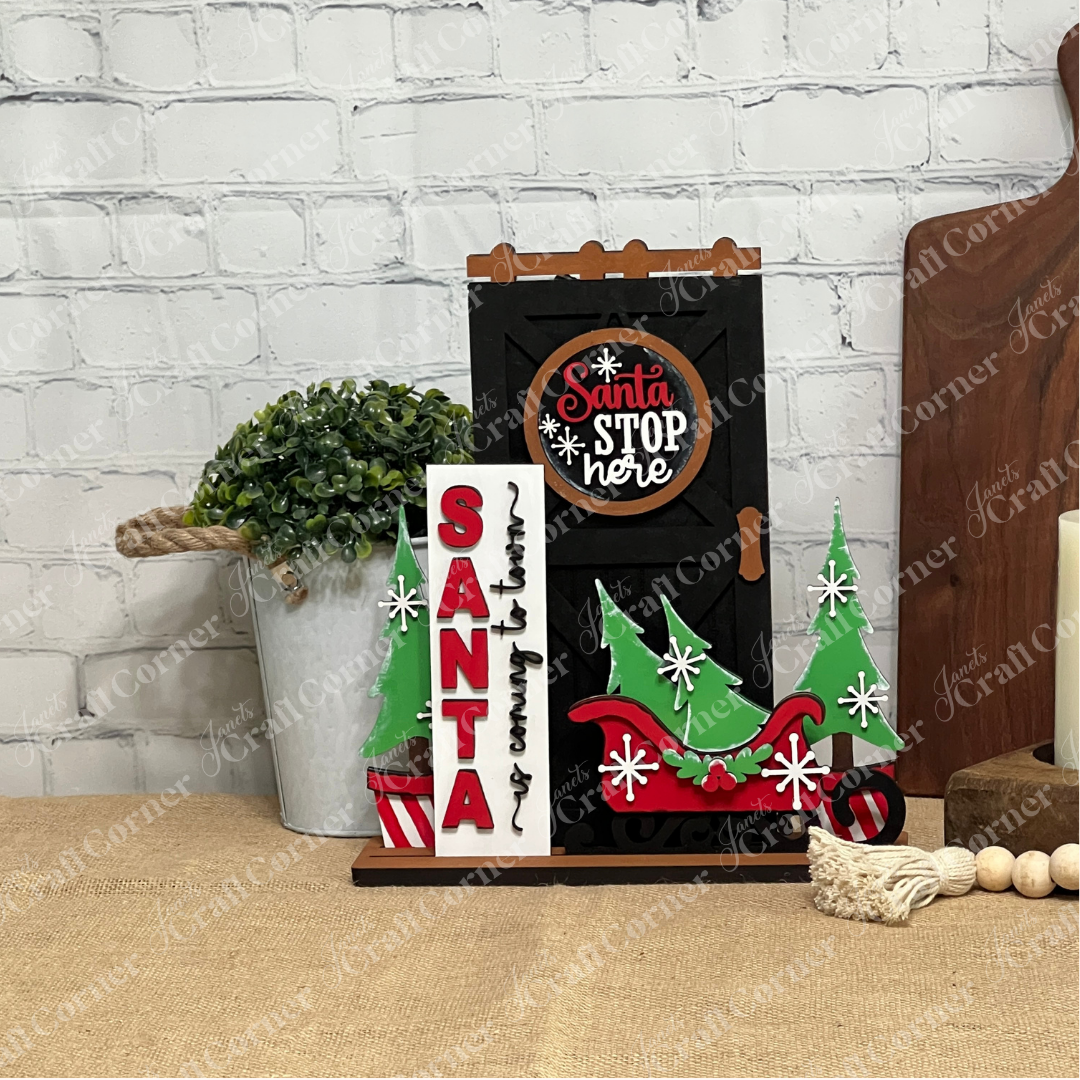 A festive display features Janet's Craft Corner’s Farmhouse Porch Interchangeable Base DIY decor kit, including a black "Santa Stop Here" sign, a red and black "Santa is coming to town" sign, and green trees with red ornaments on a wooden table.