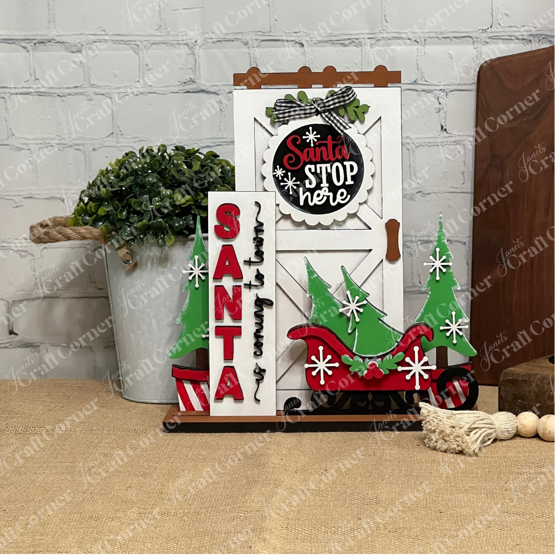 Add festive farmhouse charm to your entryway with Janet's Craft Corner Farmhouse Porch Interchangeable Base, a DIY seasonal home decor craft kit featuring a white door, "Santa Stop Here" sign, green trees, red sleigh, and more.