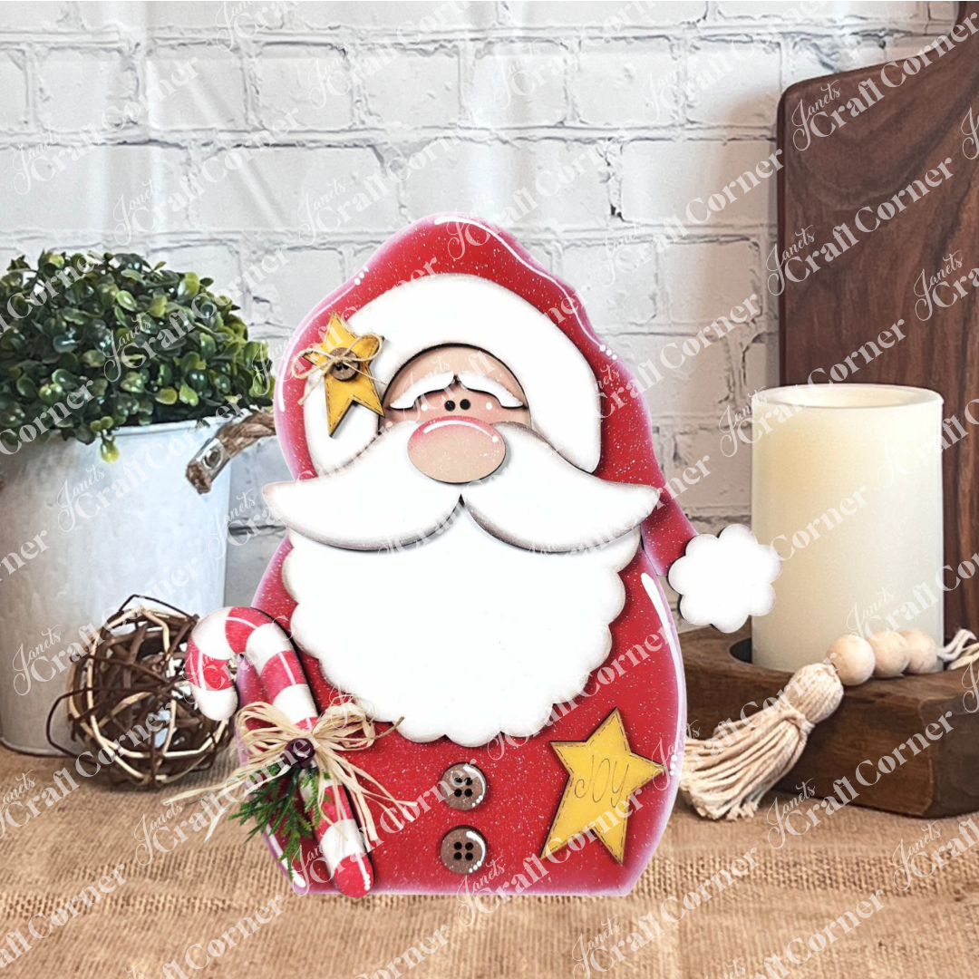 Janet's Craft Corner Santa Shelf Sitter DIY seasonal home decor craft kit (set of 1) is displayed on a wooden table with a potted plant, lit candle, wooden beads, and a white brick wall backdrop for festive charm.