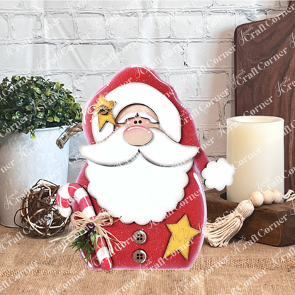 Janet's Craft Corner Santa Shelf Sitter DIY seasonal home decor craft kit (set of 1) is displayed on a wooden table with a potted plant, lit candle, wooden beads, and a white brick wall backdrop for festive charm.