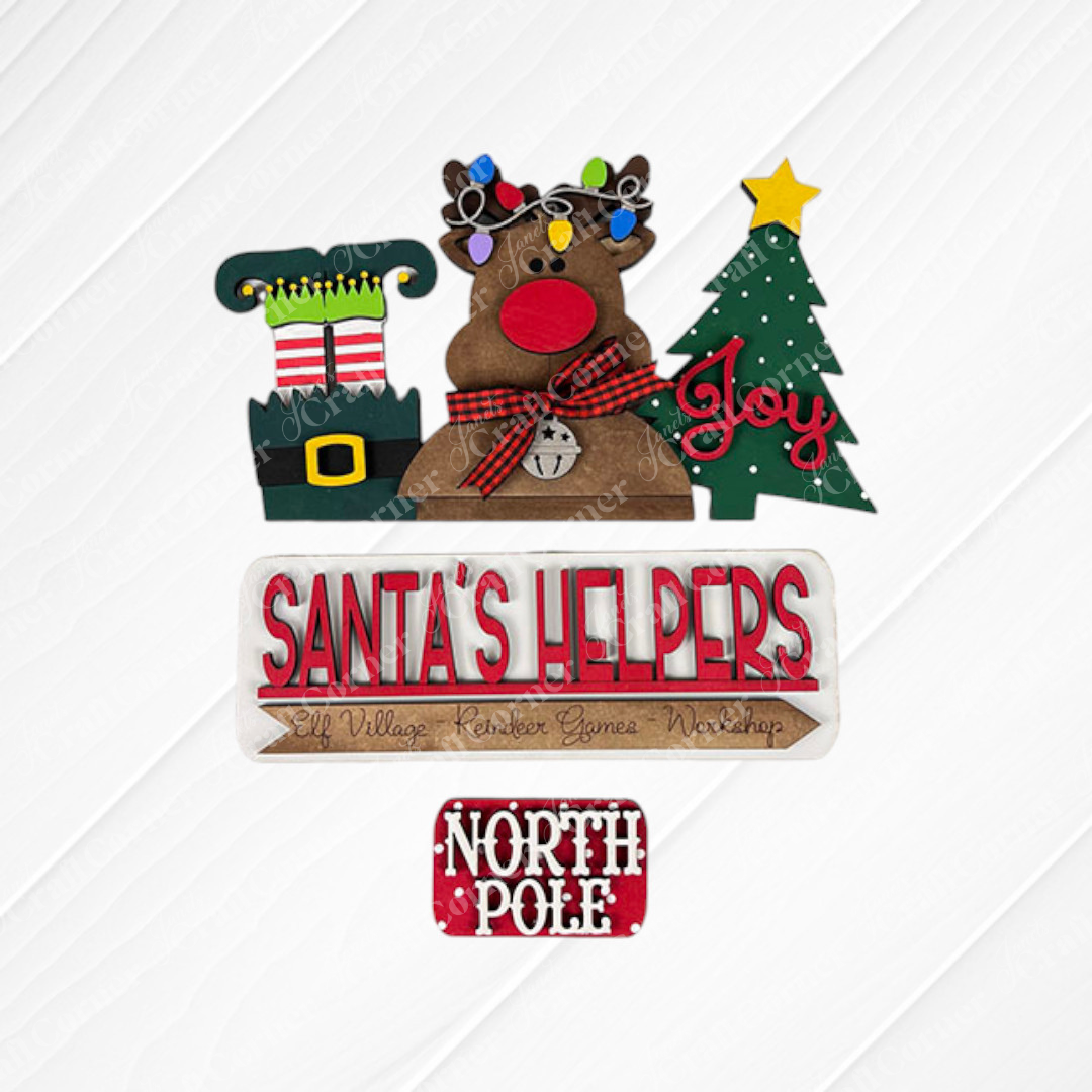 On a white surface, a festive display features a light-adorned reindeer, an elf shoe, and a green Christmas tree with the word "Joy." A sign labeled "Santa's Helpers" from Janet's Craft Corner includes an interchangeable insert for your home decor. Adding to the scene are signs that read "Elf Village Reindeer Games Workshop" and "North Pole," both crafted on burlap.