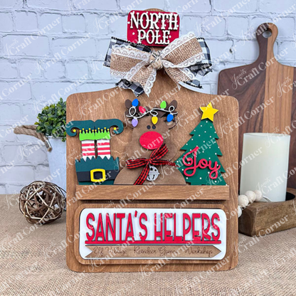 Create festive DIY decor with Janet's Craft Corner's Breadboard Interchangeable Base, featuring inserts like a reindeer with Christmas light antlers, elf hat and shoes, "Joy" tree, "SANTA'S HELPERS" text in red, topped with a ribbon-adorned "NORTH POLE" plaque.