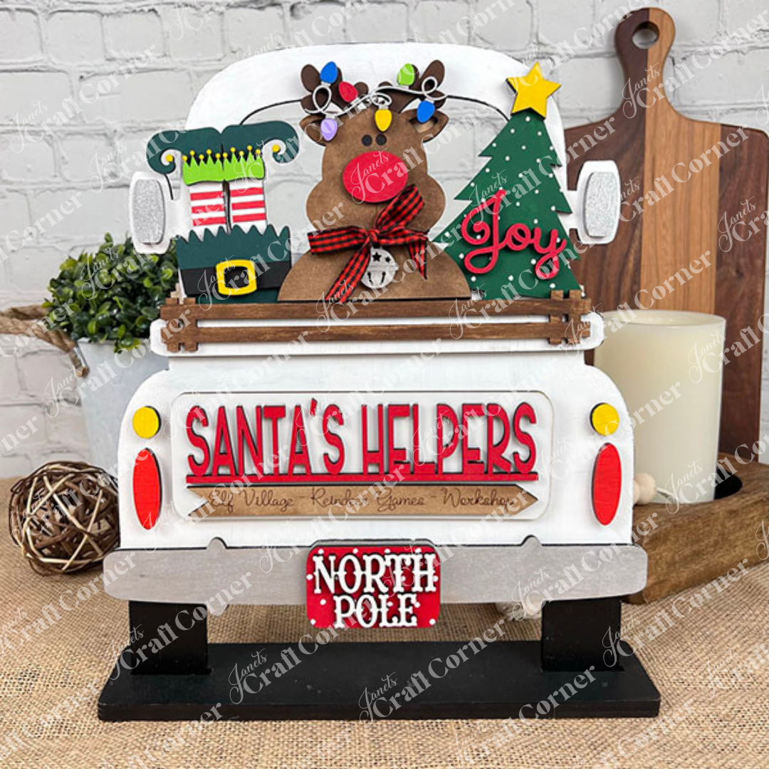A festive decoration from Janet's Craft Corner features an antique truck base with a reindeer adorned with ornaments, a Christmas tree, and gift boxes. The truck bears the phrases "SANTA'S HELPERS" and "NORTH POLE" and rests on a burlap-covered table.