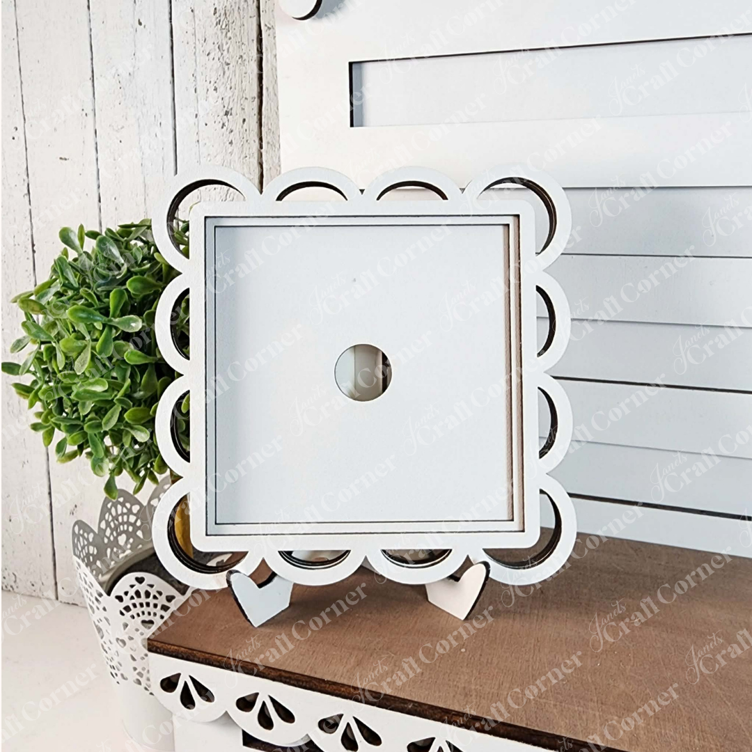 The Scallop Lace Square Frame by Janet’s Craft Corner is a white, scalloped wooden frame with a circular opening on a stand—ideal for DIY seasonal decor and displayed on wood near greenery and a decorative panel.