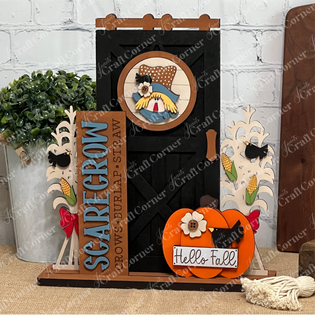 Create a charming fall display with Janet's Craft Corner Farmhouse Porch Interchangeable Base DIY kit, featuring farmhouse accents, a black barn door, scarecrow, corn stalks, “Hello Fall” pumpkin sign, and a vertical “SCARECROW” decor piece.
