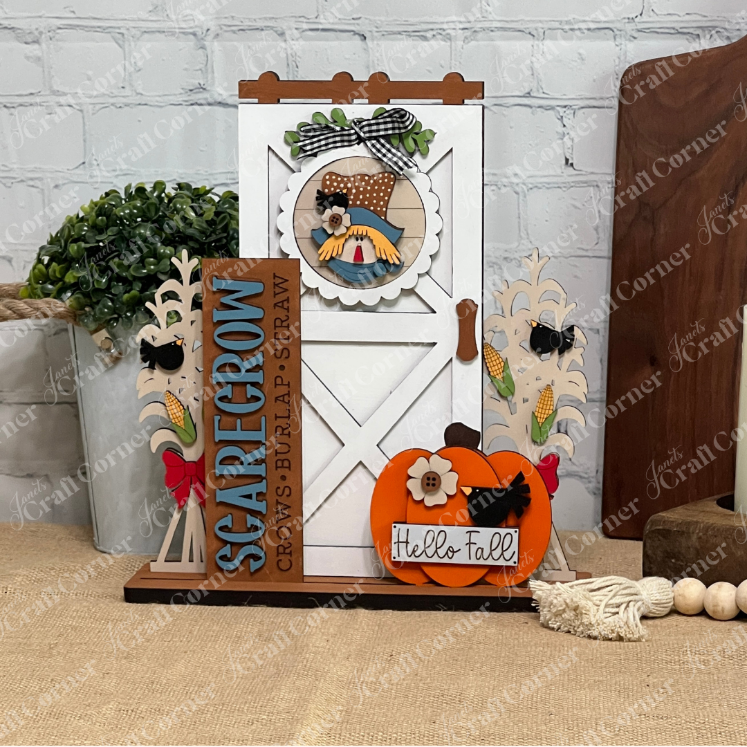 Janet's Craft Corner's Scarecrow Interchangeable Porch Set is a DIY fall decor piece with "SCARECROW," corn stalk accents, and a "Hello Fall" pumpkin on a barn door background, perfect for displaying on your table or porch.