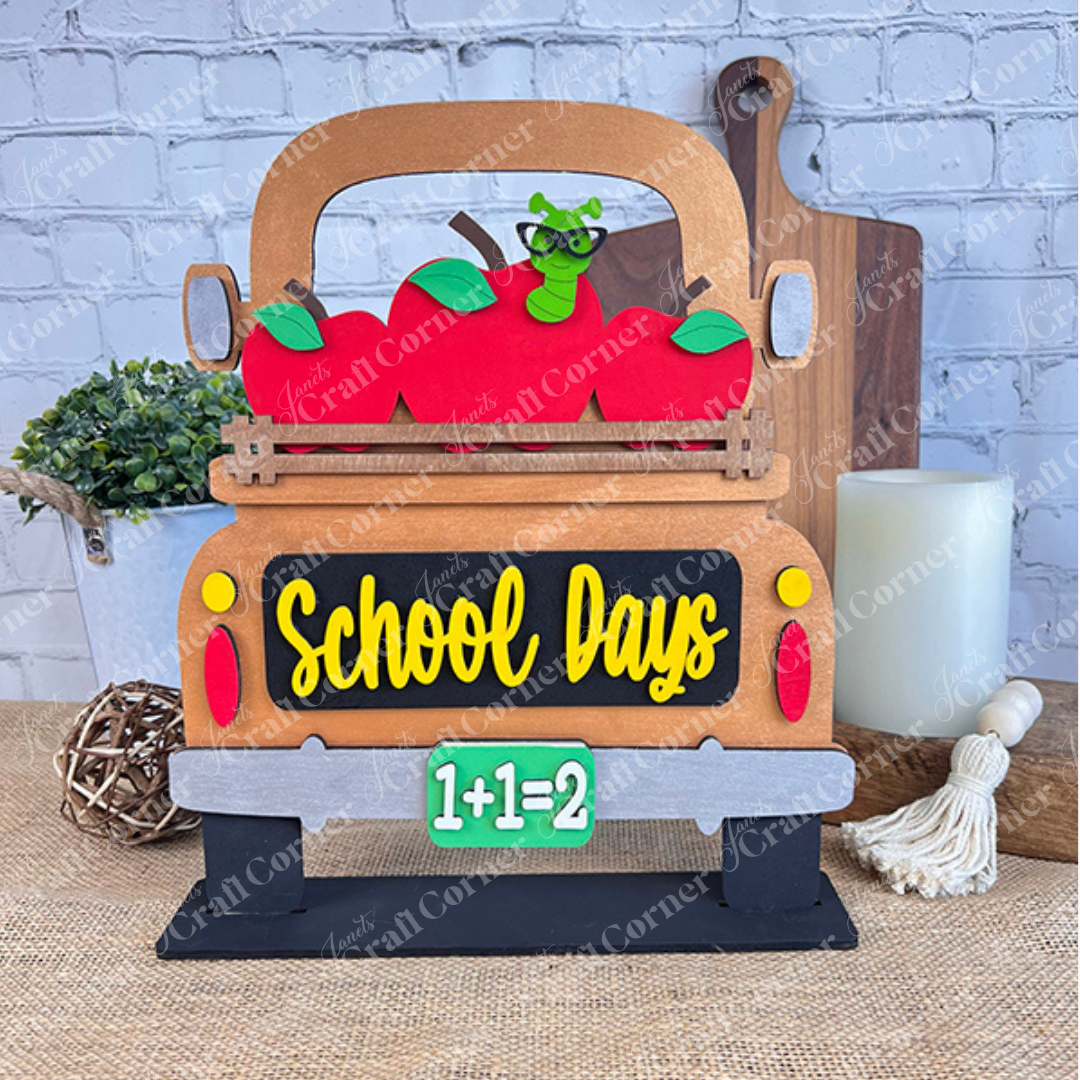 The School Days Interchangeable Insert from Janet's Craft Corner is a charming decorative piece shaped like a truck overflowing with red apples and a cartoon worm. The tailgate is adorned with the words "School Days," and the license plate reads "1+1=2." Ideal for farmhouse decor, it includes a white brick wall backdrop and various whimsical accents.