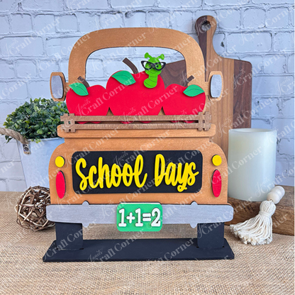 The School Days Interchangeable Insert from Janet's Craft Corner is a charming decorative piece shaped like a truck overflowing with red apples and a cartoon worm. The tailgate is adorned with the words "School Days," and the license plate reads "1+1=2." Ideal for farmhouse decor, it includes a white brick wall backdrop and various whimsical accents.