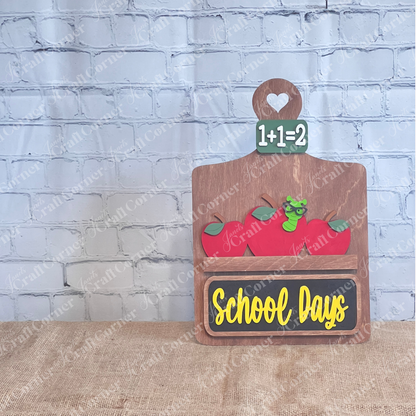 Featuring three red apples and a cartoon green worm with glasses above the words "School Days" in yellow, the School Days Interchangeable Insert by Janet's Craft Corner is perfect for farmhouse decor. This DIY home decor kit is topped with a gingham and burlap bow, beautifully set against a brick wall background.