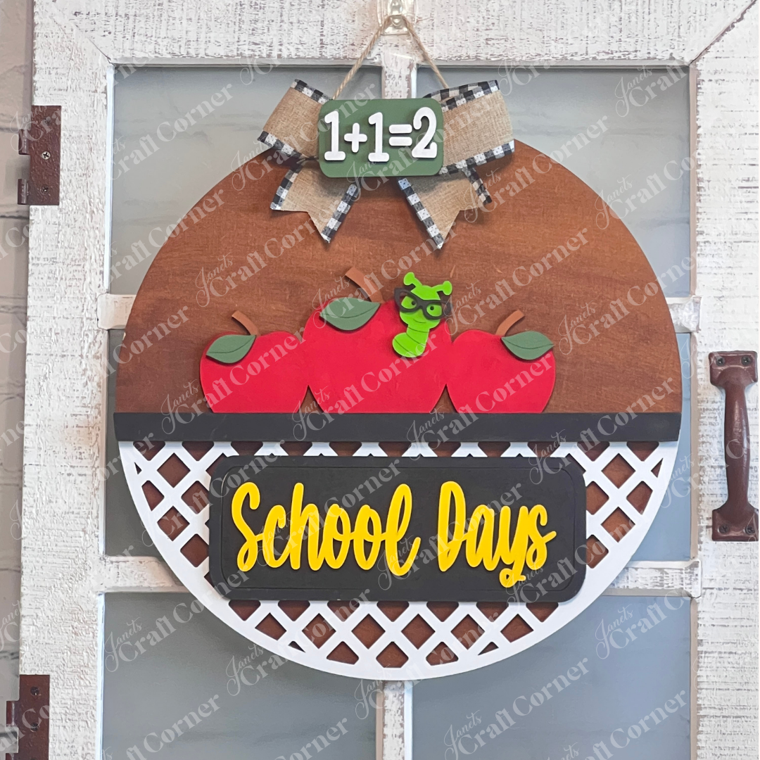 Against a white door window back drop Janet's Craft Corner's School Days Interchangeable Insert features a worm with three red apples and "School Days" in yellow. The DIY kit includes a small green chalkboard with "1+1=2" and a bow—ideal for interchangeable home decor.