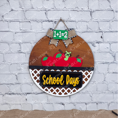 Against a white brick backdrop, Janet's Craft Corner's School Days Interchangeable Insert features a worm with three red apples and "School Days" in yellow. The DIY kit includes a small green chalkboard with "1+1=2" and a bow—ideal for interchangeable home decor.