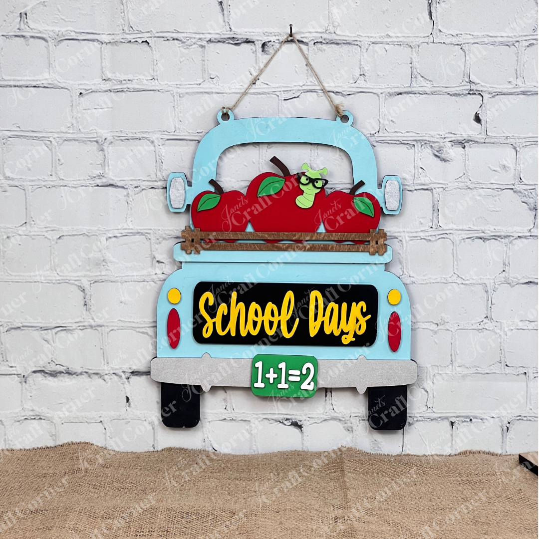 The School Days Interchangeable Insert by Janet's Craft Corner is a DIY decor kit featuring a light blue truck with red apples and green leaves. It displays "School Days" in yellow and "1+1=2" in green on black, perfect for farmhouse decor, adding charm against any white brick wall.