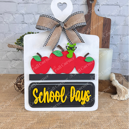 The School Days Interchangeable Insert by Janet's Craft Corner is a charming DIY home decor kit featuring a decorative sign adorned with three red apples and an adorable green worm wearing glasses. A black plaque below displays the words "School Days" in yellow, and it is completed with a black and white checkered bow accented with burlap, adding a touch of farmhouse decor charm.