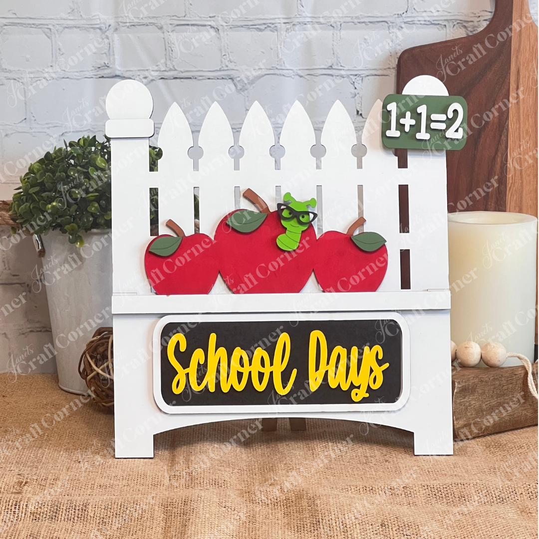 The School Days Interchangeable Insert by Janet's Craft Corner features a white picket fence with red apple cutouts and a worm, adorned with "1+1=2" and "School Days" on a green board and blackboard. Set against burlap and brick, it brings quaint farmhouse charm to any space.