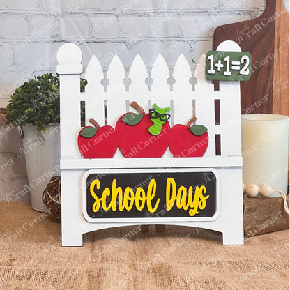 The School Days Interchangeable Insert by Janet's Craft Corner features a white picket fence with red apple cutouts and a worm, adorned with "1+1=2" and "School Days" on a green board and blackboard. Set against burlap and brick, it brings quaint farmhouse charm to any space.
