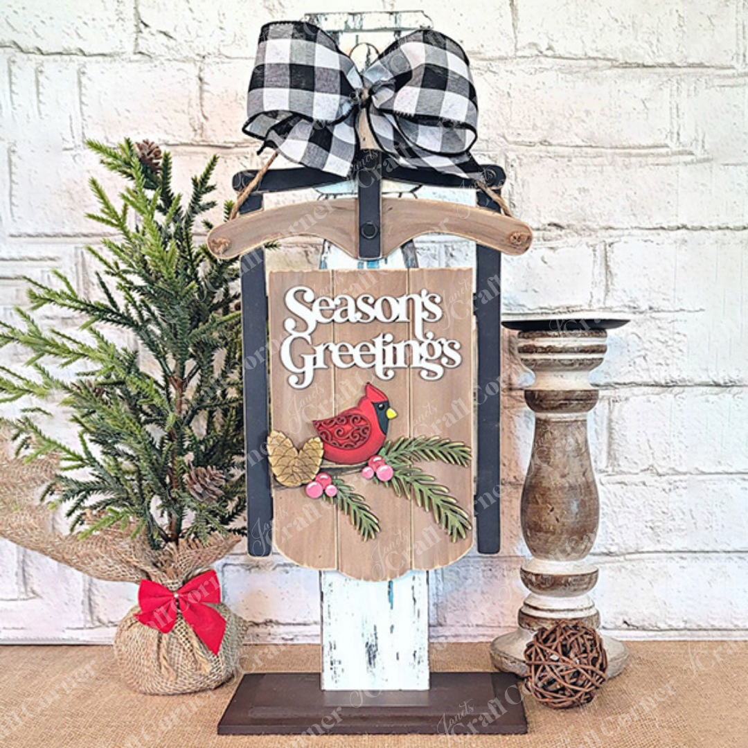 Create a festive holiday atmosphere with the Season's Greetings Sled Door Hanger from Janet's Craft Corner, featuring a red cardinal and pinecones. Adorned with a black-and-white checkered bow on top, this charming display includes a small burlap-wrapped pine tree on the left and a wooden candle holder on the right – perfect for your own DIY projects or as part of your seasonal decor.