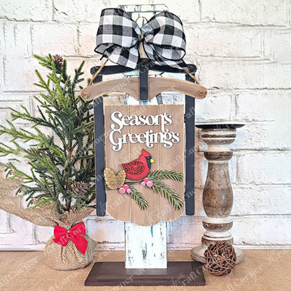 Create a festive holiday atmosphere with the Season's Greetings Sled Door Hanger from Janet's Craft Corner, featuring a red cardinal and pinecones. Adorned with a black-and-white checkered bow on top, this charming display includes a small burlap-wrapped pine tree on the left and a wooden candle holder on the right – perfect for your own DIY projects or as part of your seasonal decor.