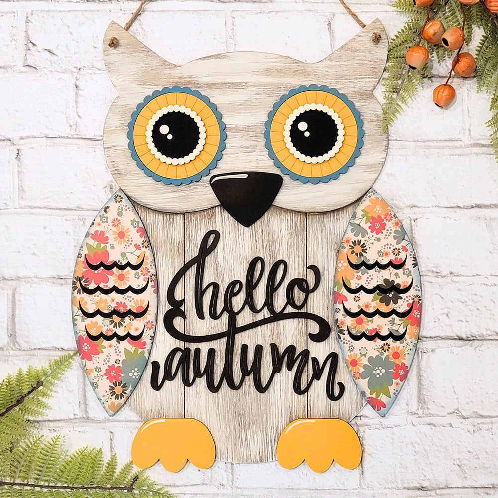 The Shiplap Owl Door Hanger from Janet's Craft Corner features a charming design with large colorful eyes and wings adorned in floral patterns, showcasing the message "Hello Autumn" in an elegant script. Set against a white brick backdrop, this decorative piece enhances your DIY home decor with fall-themed greenery and small orange berries.