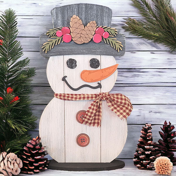 Introducing the Shiplap Snowman Shelf Sitter from Janet's Craft Corner: This charming piece features a snowman with a plaid scarf and buttons, topped with a hat adorned with pinecones and berries. It is beautifully surrounded by pinecones and evergreen branches, all set against a rustic wooden plank backdrop. A delightful addition to your home décor!