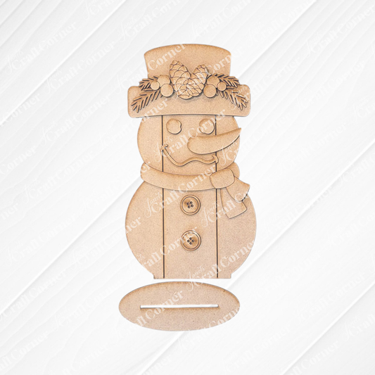 Introducing Janet's Craft Corner Shiplap Snowman Shelf Sitter, a DIY seasonal home decor kit. This wooden cutout snowman is adorned with pinecone and leaf embellishments, featuring a charming hat, scarf, buttons, and classic carrot nose on a textured wooden backdrop.