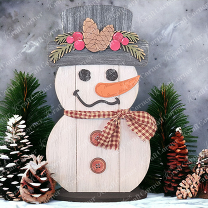 Introducing the Shiplap Snowman Shelf Sitter from Janet's Craft Corner: This charming piece features a snowman with a plaid scarf and buttons, topped with a hat adorned with pinecones and berries. It is beautifully surrounded by pinecones and evergreen branches, all set against a concrete backdrop. A delightful addition to your home décor!