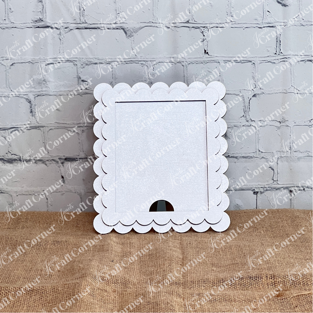 Janet's Craft Corner Single Scallop Trim Farmhouse Frame in white stands upright on burlap, featuring a cut-out bottom for interchangeable display, with a light gray brick wall as the background.