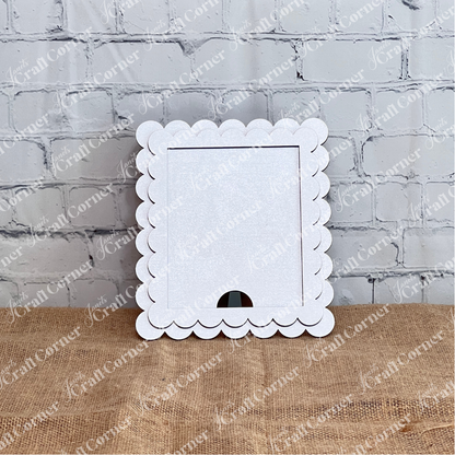 Janet's Craft Corner Single Scallop Trim Farmhouse Frame in white stands upright on burlap, featuring a cut-out bottom for interchangeable display, with a light gray brick wall as the background.