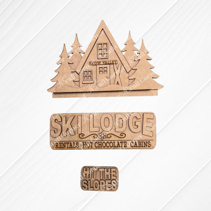Janet's Craft Corner Ski Lodge Interchangeable Insert DIY craft kit: 3 laser-cut wood pieces—a triangle ski cabin with "Snow Valley," a "Ski Lodge Rentals • Hot Chocolate • Cabins" sign, and a "HIT THE SLOPES" sign.