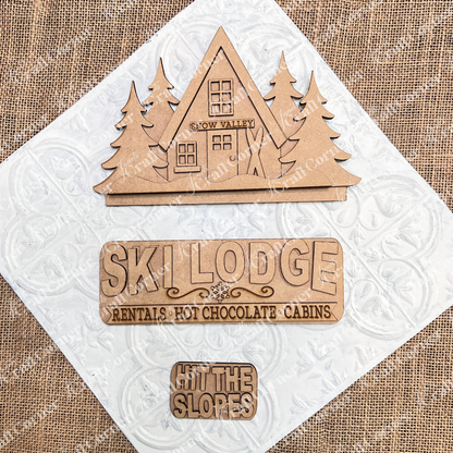 Janet's Craft Corner Ski Lodge Interchangeable Insert DIY craft kit: 3 laser-cut wood pieces—a triangle ski cabin with "Snow Valley," a "Ski Lodge Rentals • Hot Chocolate • Cabins" sign, and a "HIT THE SLOPES" sign.