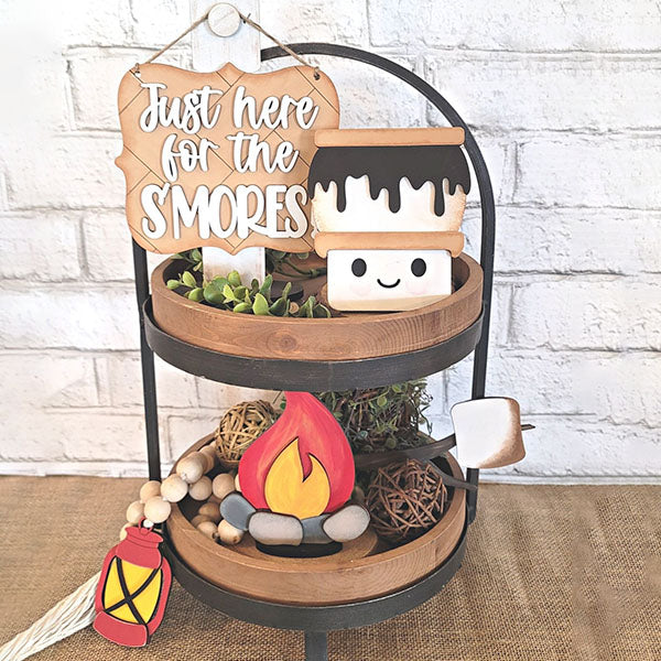 A tiered display features a delightful "Just here for the S'mores" sign alongside Janet's Craft Corner's S'mores - Tiered Tray Kit. This kit includes an adorable marshmallow character, a decorative campfire, and DIY elements such as a marshmallow on a stick and a lantern ornament, brilliantly enhancing any home décor when placed on its wooden tabletop.