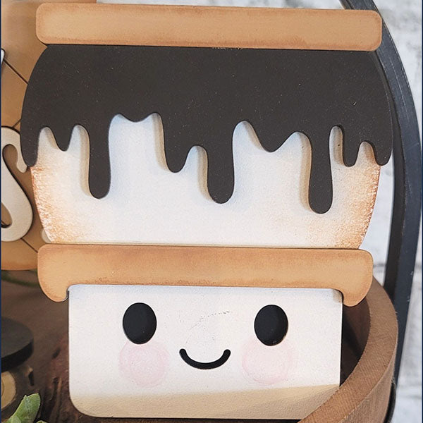 The S'mores - Tiered Tray Kit from Janet's Craft Corner features an adorable cartoon s'more character with a smiling face as its main attraction. The design includes a layer of melted chocolate drizzling over a toasted marshmallow, all perfectly nestled between two graham crackers, making it a charming addition to your home décor.