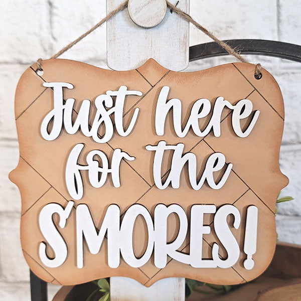 A decorative sign from Janet's Craft Corner's S'mores - Tiered Tray Kit hangs elegantly from a string, ideal for enhancing your tiered tray display. It showcases the phrase "Just here for the S'MORES!" in stylish white lettering on a brown, wood-like backdrop. The wavy edge of this home décor piece beautifully complements any light wall.