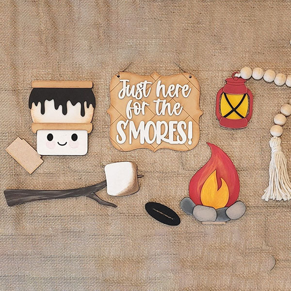 Introducing the S'mores Tiered Tray Kit from Janet's Craft Corner, featuring charming s'mores-themed items on burlap. This delightful DIY kit includes a smiling marshmallow, a graham cracker, chocolate piece, a wooden sign proclaiming "Just here for the S'MORES!", a campfire replica, lantern, and beaded garland with tassels. Perfect for enhancing your home décor!