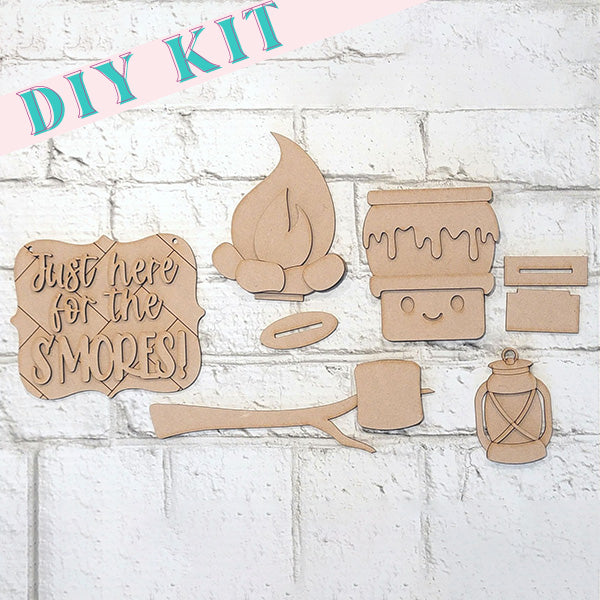 Introducing Janet's Craft Corner's S'mores - Tiered Tray Kit, a delightful DIY kit featuring unfinished wood cutouts against a white brick background. This charming set includes pieces resembling a sign that reads "Just here for the s'mores!", along with a marshmallow on a stick, a campfire, a jar, and a lantern—perfect for cozy home décor.