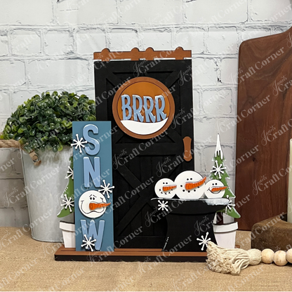 The Snow Interchangeable Porch Set by Janet's Craft Corner is a winter-themed unfinished wood kit with a black door, orange "BRRR" sign, snowmen, pine trees, and a blue "SNOW" sign—perfect DIY home decor for the season.