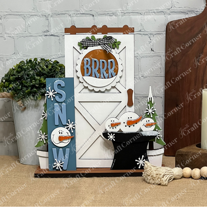 Janet's Craft Corner Snow Interchangeable Porch Set: an unfinished wood kit with snowmen, snowflakes, pine trees, and "BRRR" and "SNW" on a barn-style backdrop in neutral and blue—ideal rustic winter DIY home decor.