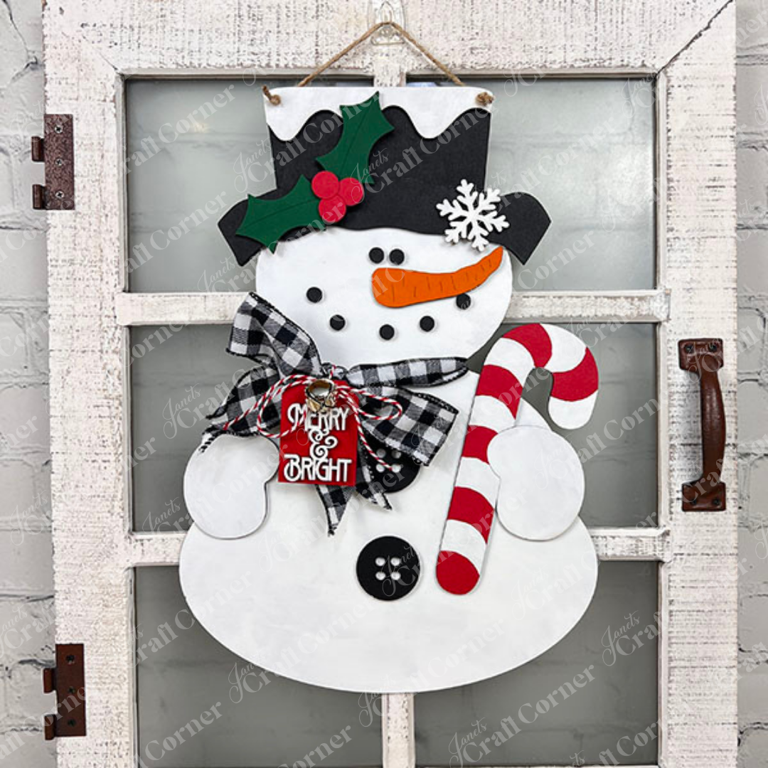 Decorate your home with the Snowman Door Hanger from Janet's Craft Corner, featuring a festive snowman on a rustic window pane backdrop. This charming door hanger showcases a black hat adorned with holly, a plaid scarf, and holds a candy cane. A small "Merry & Bright" sign adds an extra touch of cheer to its chest. It's perfect for enhancing your décor against any brick wall background.