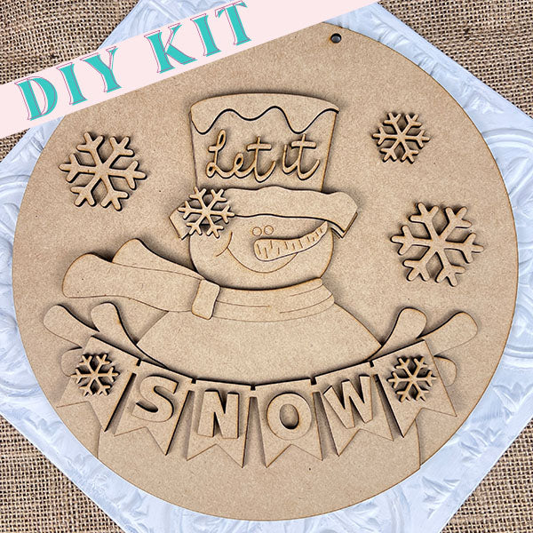 Janet's Craft Corner presents the "Snowman Let It Snow Door Hanger" DIY kit, showcasing a cheerful snowman with the words "let it" on its hat and surrounded by delicate snowflakes. Below, a banner reads "SNOW," adorned with small snowflakes on either side. Ideal for holiday decorations or as an enchanting door hanger, this kit is set against a textured background.