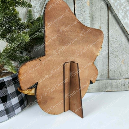 The Snowman Face Shelf Sitter from Janet’s Craft Corner—a DIY home decor craft kit—rests on a small easel beside a potted evergreen in a plaid container, with a rustic wooden backdrop behind them, making for an inviting piece of winter home decor.
