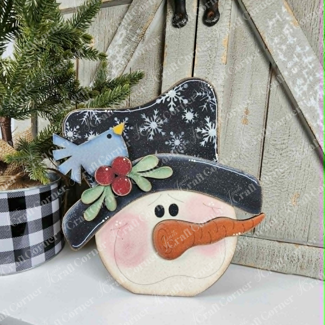 Add a cheerful winter touch to your home with the Snowman Face Shelf Sitter from Janet’s Craft Corner—a DIY home decor craft kit featuring a smiling snowman with a carrot nose, wearing a black hat decorated with snowflakes, holly, and a cute blue bird. The kit also includes a small pine tree in a plaid pot, all designed to sit together against a rustic wooden door background for charming seasonal decor. Set of 1.