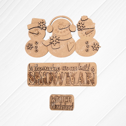 On a textured surface, the Snowman Interchangeable Insert from Janet's Craft Corner showcases wooden snowmen and text decorations with messages such as "In the meadow we can build a SNOWMAN" and "WINTER Wonderland." This hand-painted DIY home decor kit includes snowmen adorned with scarves, hats, and snowflake decorations for unique interchangeable home accents.