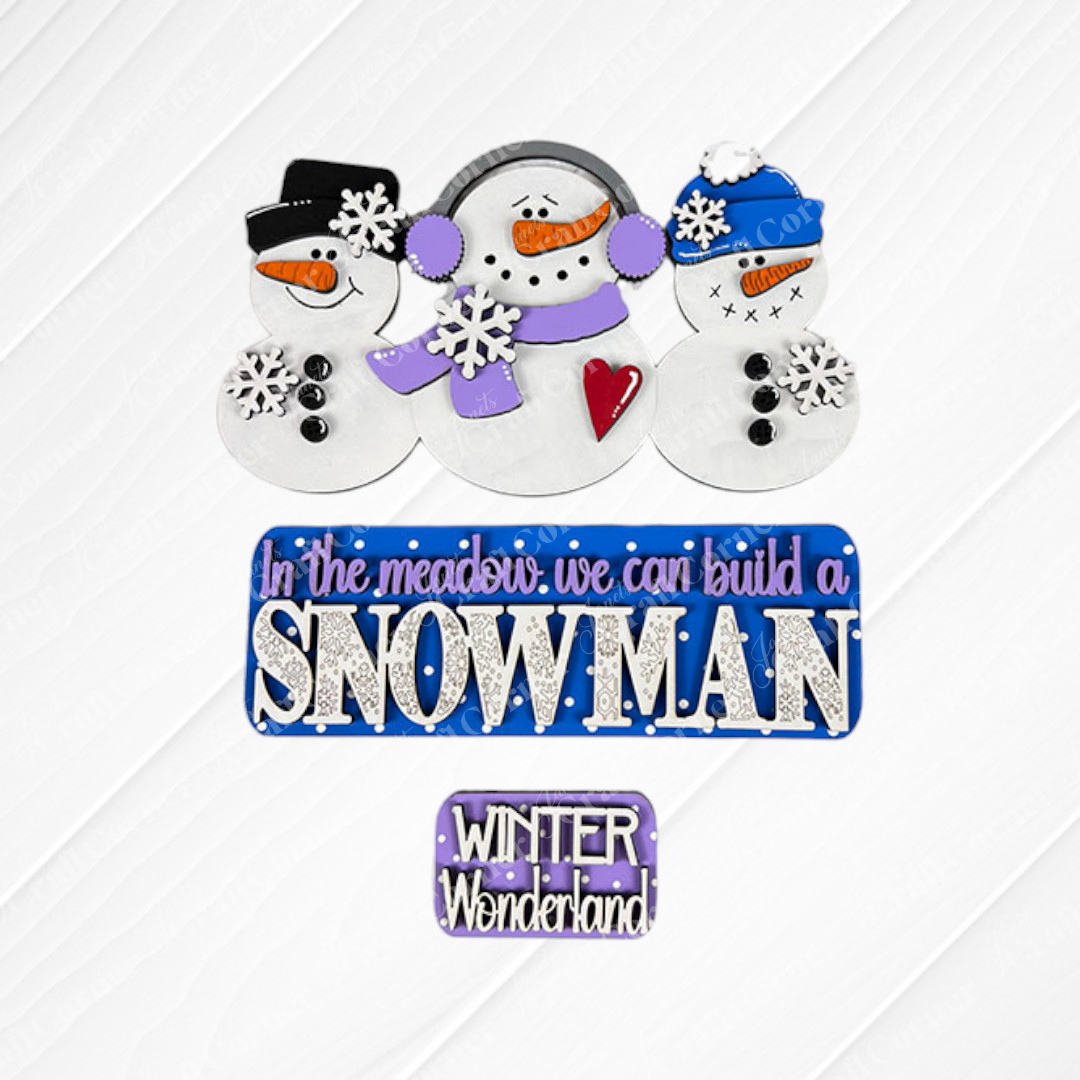 Using the Snowman Interchangeable Insert DIY home decor kit from Janet's Craft Corner, three snowmen adorned with hats and scarves stand against a decorative snowy background. A sign reading "In the meadow, we can build a SNOWMAN" is depicted in purple and blue letters, while below it, a smaller sign declares "WINTER Wonderland." This scene is beautifully accented with snowflakes for an interchangeable home decor feel.