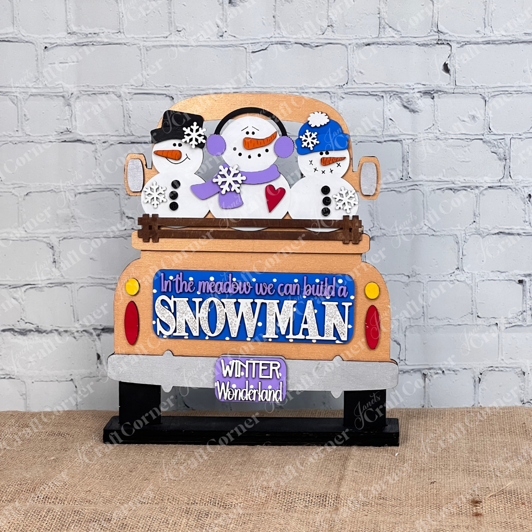 The Snowman Interchangeable Insert by Janet's Craft Corner is a DIY home decor kit featuring a wooden piece with three snowmen in a truck and a blue sign that says "In the meadow we can build a snowman" and license plate "Winter Wonderland," plus a snowman insert against a white brick wall.