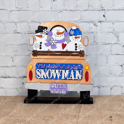 The Snowman Interchangeable Insert by Janet's Craft Corner is a DIY home decor kit featuring a wooden piece with three snowmen in a truck and a blue sign that says "In the meadow we can build a snowman" and license plate "Winter Wonderland," plus a snowman insert against a white brick wall.