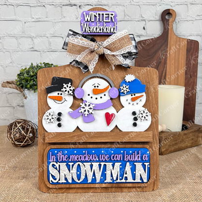 Janet's Craft Corner offers a Breadboard Interchangeable Base with Insert DIY kit. The base features three snowmen: one in a black hat, one in a purple scarf, and one in a blue hat. Signs read "Winter Wonderland" and "In the meadow we can build a snowman," making it perfect for festive home decor.