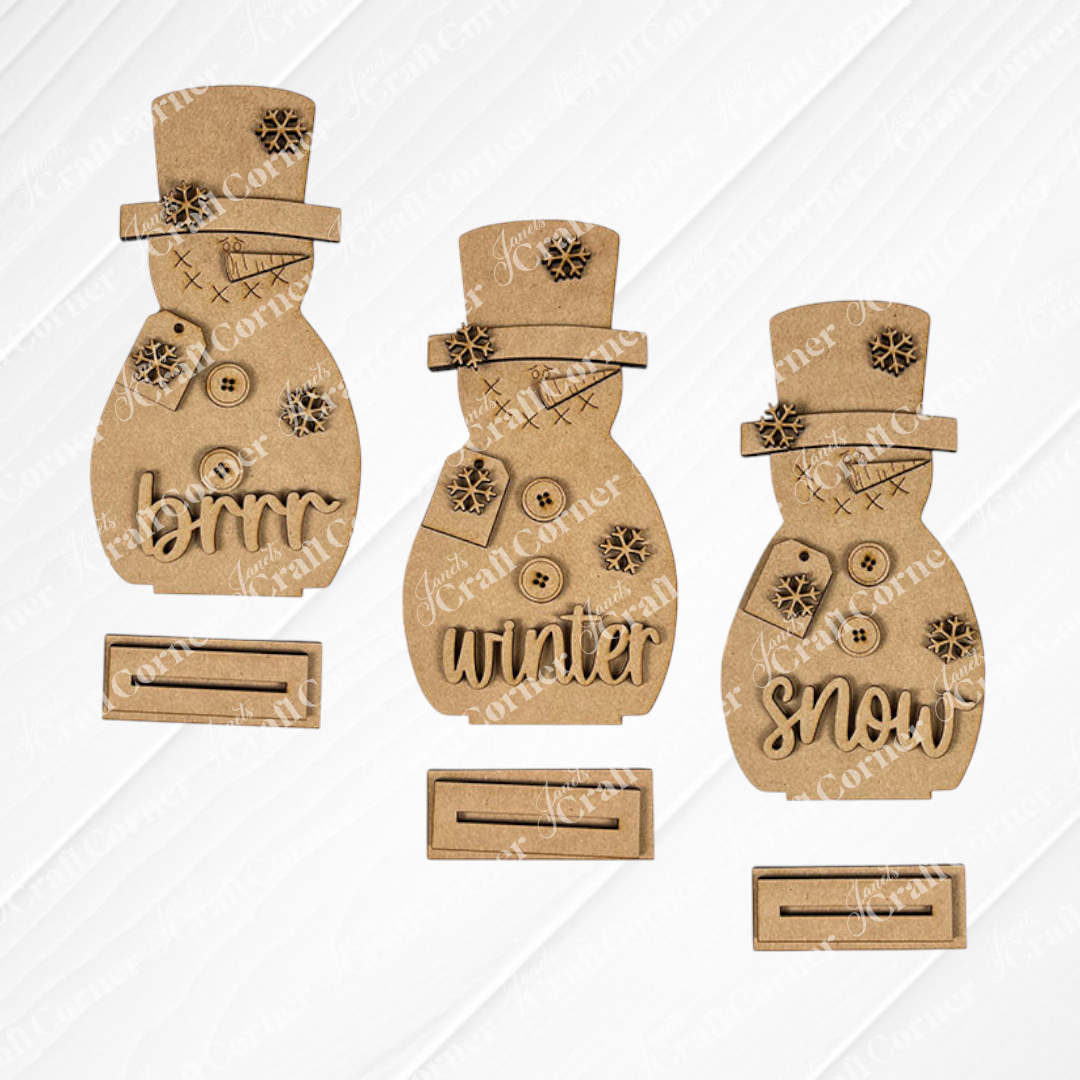 Janet's Craft Corner Snowman Shelf Sitters—three unfinished cardboard snowmen with buttons and "snow," "brrr," and "winter" accessories—are displayed on a white textured surface with stands, perfect for winter décor.