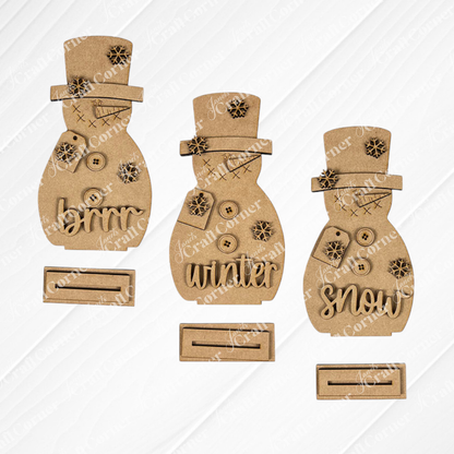 Janet's Craft Corner Snowman Shelf Sitters—three unfinished cardboard snowmen with buttons and "snow," "brrr," and "winter" accessories—are displayed on a white textured surface with stands, perfect for winter décor.
