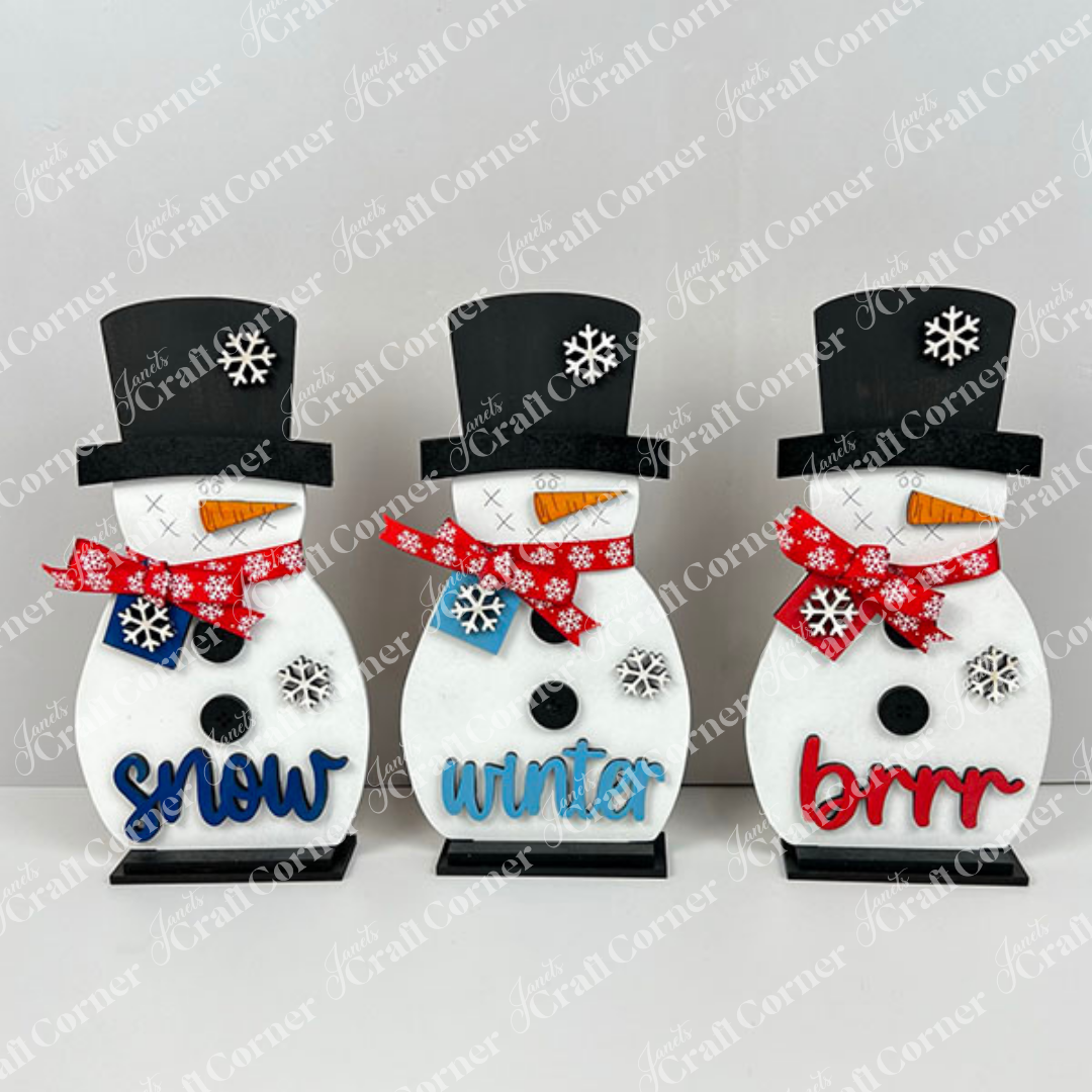 Janet's Craft Corner Snowman Shelf Sitters feature three snowmen in black hats and red or blue scarves, each with a snowflake design and festive word—ideal for winter or holiday home decor.